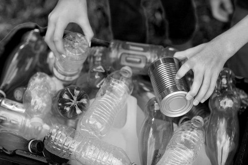 A pair of hands sorting and handling recyclable waste such as plastic bottles and tin cans over a collection of various empty plastic bottles and metal cans, some with labels and different shapes, in a rubbish disposal area outside on a flat surface. The scene appears to be part of a waste clearance process, possibly in the vicinity of Stoke Newington, where Commercial Waste Stoke Newington operates rubbish removal services. The background is blurred, focusing the viewer's attention on the waste items and the manual sorting activity. The collection includes clear plastic bottles, some with caps, and a metal tin can, with textures ranging from smooth plastic to shiny, reflective metal surfaces. The image is in black and white, emphasizing the shapes and materials of the waste objects. The environment suggests an outdoor space used for waste collection, aligned with professional rubbish clearance and recycling efforts typical of the area.