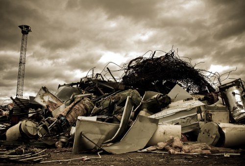 A large pile of discarded metal and household appliances, including washing machines, refrigerators, and broken furniture, is stacked outdoors on a paved surface, likely part of a rubbish removal or clearance site in Stoke Newington. The metallic objects display signs of rust and corrosion, with some parts bent or damaged. Behind the pile, there is a dark, tangled mass of wires and debris, indicating an accumulation of scrap material. To the left, a tall metallic tower or pole stands against a cloudy sky with hints of sunlight breaking through, casting a muted light over the scene. This image reflects the type of waste that Commercial Waste Stoke Newington might handle during clearance services, situated within a typical urban environment close to residential or commercial premises, emphasizing the need for proper disposal or recycling of bulky household and industrial waste.