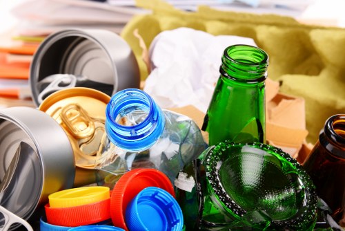 A close-up view of a collection of clean recyclable bottles and containers made of glass and plastic, including a green glass bottle and a clear plastic water bottle with a blue cap, surrounded by colorful plastic bottle caps in red, yellow, and blue. These items are placed on a surface outdoors, with a background that features cardboard boxes and yellow waste packaging material, indicating a recycling or rubbish collection scene possibly related to waste clearance services in Stoke Newington. The image emphasizes the presence of glass and plastic waste materials, commonly handled by commercial waste removal companies like Commercial Waste Stoke Newington, situated in the N16 postcode area, and is suitable for illustrating rubbish disposal or recycling services in the local context.