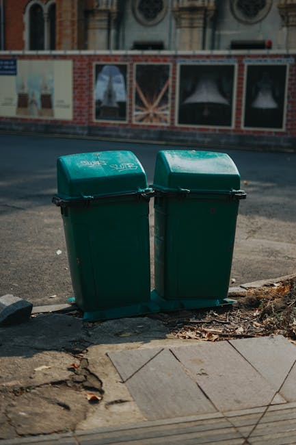 Two green plastic rubbish bins with hinged lids are positioned side by side on a paved sidewalk, close to the edge of a street. The bins are made of textured, durable plastic and are slightly weathere